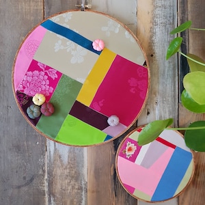 May include: Two circular embroidery hoops with colourful fabric patchwork designs. The larger hoop has a pink, yellow, green, and brown patchwork design. The smaller hoop has a pink, blue, and white patchwork design.