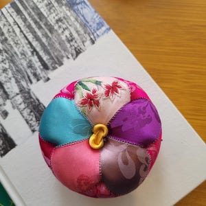 May include: A colorful, round pincushion with a gold-colored clasp. The pincushion is made of fabric patches in various colors, including pink, teal, purple, and brown. Some patches feature floral patterns. The pincushion is resting on a white surface.