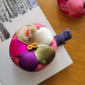May include: A colorful, round fabric ornament with a gold clasp. The ornament is made of various fabric patches in shades of pink, purple, green, and beige, with a floral embroidered design. A purple fabric strip is attached to the side. The ornament is resting on a white surface.