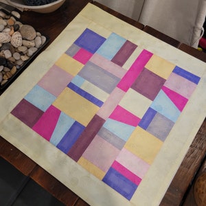 May include: A light yellow square cushion with a geometric pattern of pink, purple, blue, and yellow squares. The cushion is made of a soft, textured fabric.