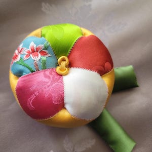 May include: A colorful, round fabric object with a yellow center and segments of red, green, pink, white, and blue. The blue segment features floral embroidery. A yellow loop and green ribbon are attached. The object is likely a decorative item or toy.