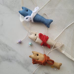 May include: Three handmade fabric fish ornaments with different coloured fabric and string accents. The fish are hanging from a white string.