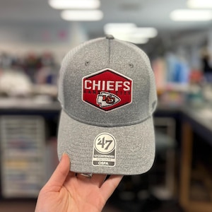 May include: A gray baseball cap with a curved brim. The cap features a red and white Chiefs logo on the front, with the word "CHIEFS" above the logo. The cap also has a "47" tag.