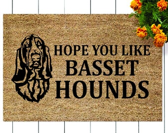 May include: A brown coir doormat with the text "HOPE YOU LIKE BASSET HOUNDS" in black lettering. A black silhouette of a Basset Hound dog is to the left of the text.