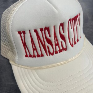 May include: A white trucker hat with the embroidered text "KANSAS CITY" in red. The hat features a cream-colored brim and a mesh back, with a cream-colored rope detail above the brim.