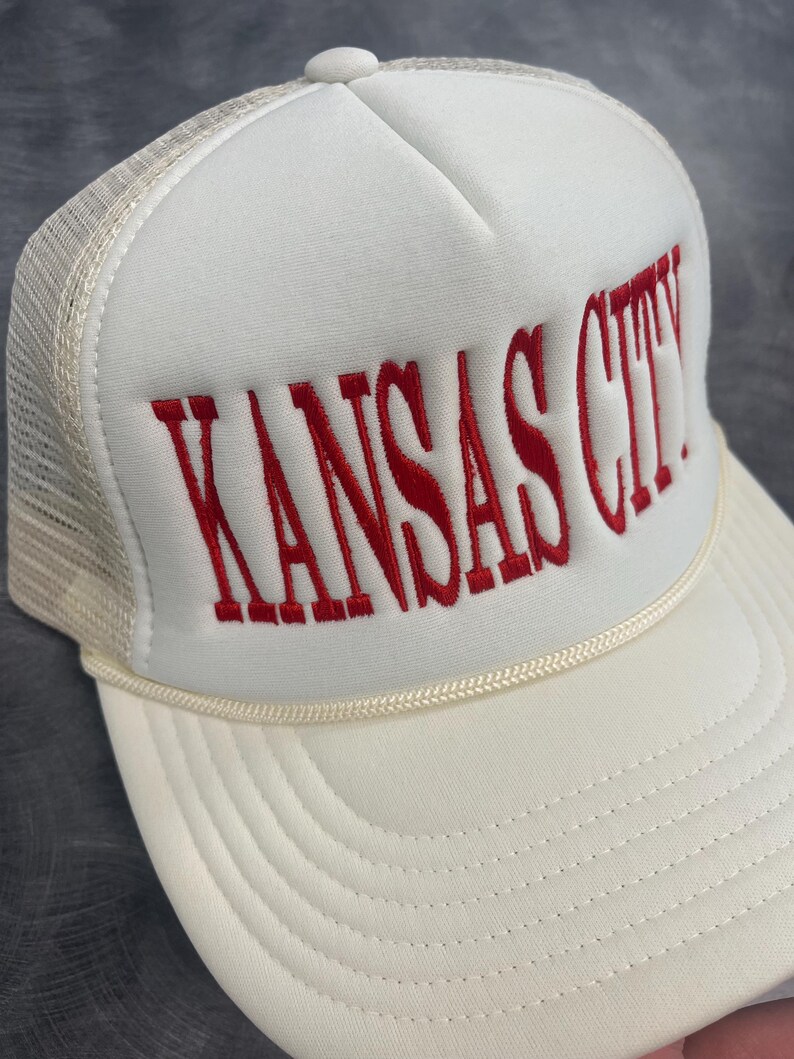 May include: A white trucker hat with the embroidered text "KANSAS CITY" in red. The hat features a cream-colored brim and a mesh back, with a cream-colored rope detail above the brim.