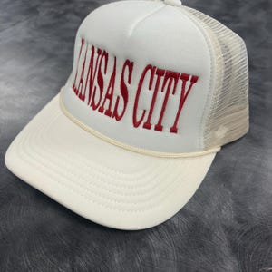 May include: A white trucker hat with the embroidered text "KANSAS CITY" in red. The hat features a cream-colored brim and a mesh back panel. A cream-colored rope detail accents the brim.