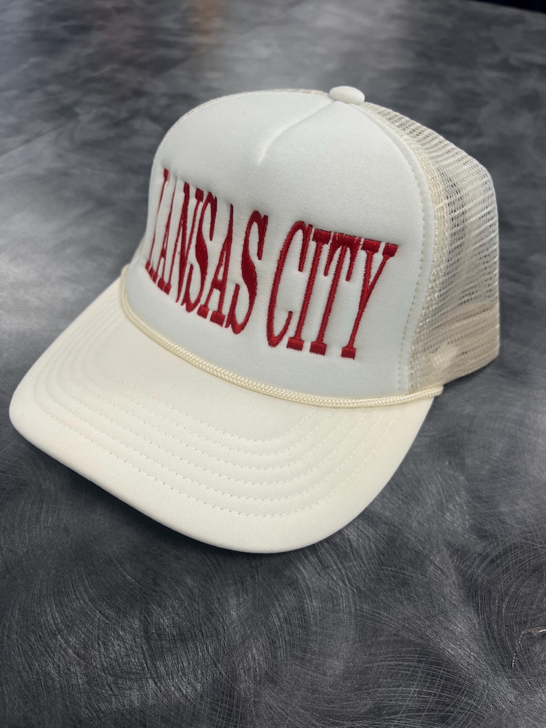 May include: A white trucker hat with the embroidered text "KANSAS CITY" in red. The hat features a cream-colored brim and a mesh back panel. A cream-colored rope detail accents the brim.