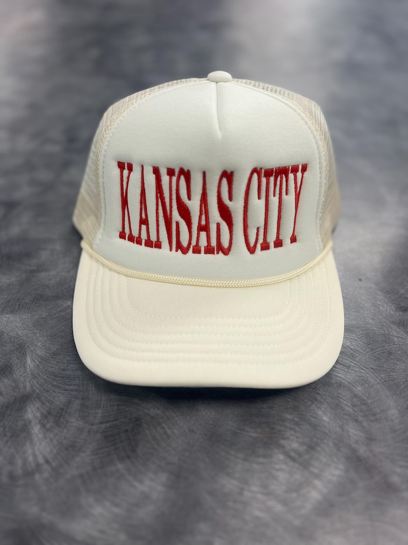 May include: Cream-colored trucker hat with the embroidered text "KANSAS CITY" in red. The hat features a white front panel, a beige mesh back, and a curved brim. A cream-colored rope detail accents the brim.