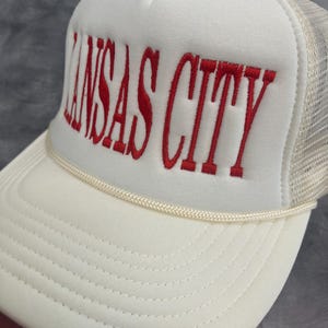 May include: A white trucker hat with the embroidered text "KANSAS CITY" in red. The hat features a mesh back and a curved brim, with a cream-colored rope detail along the front.