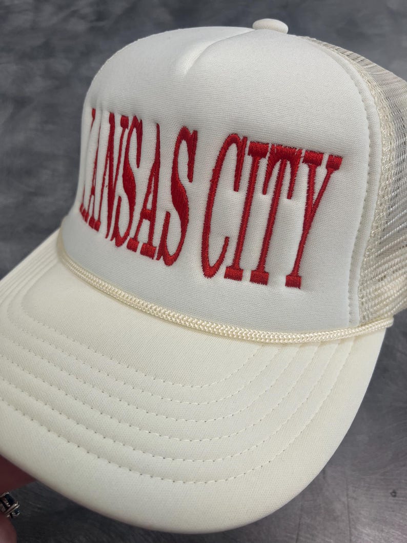 May include: A white trucker hat with the embroidered text "KANSAS CITY" in red. The hat features a mesh back and a curved brim, with a cream-colored rope detail along the front.