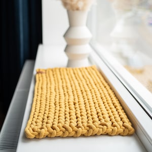 K&ouml;nnte beinhalten: Senfgelber rechteckiger Strickteppich. Der Teppich besteht aus dickem, strukturiertem Garn und hat einen geflochtenen Rand. Im Hintergrund steht eine wei&szlig;e Vase mit getrocknetem Pampasgras. Der Teppich ist ca. 61 cm lang und 41 cm breit.