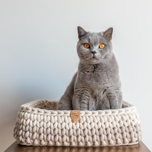 May include: A gray cat with bright yellow eyes sits in a cream-colored, hand-knitted cat bed. The bed is rectangular and has a woven texture. A small, brown leather tag is attached to the front of the bed.