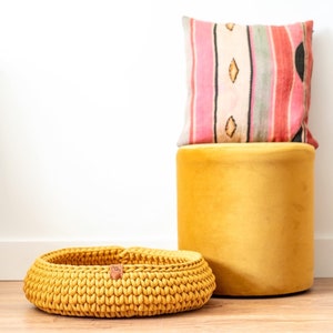 May include: A mustard yellow, hand-knitted basket with a leather tag, a matching yellow velvet ottoman, and a decorative pillow with a striped pattern in pink, red, and cream colors.