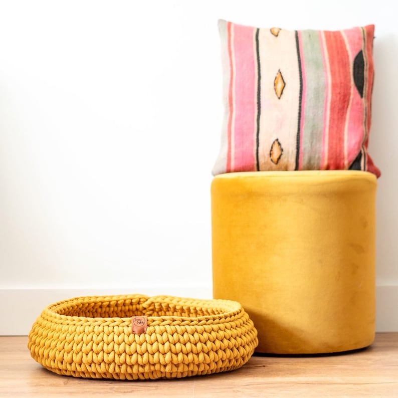 May include: A mustard yellow, hand-knitted basket with a leather tag, a matching yellow velvet ottoman, and a decorative pillow with a striped pattern in pink, red, and cream colors.