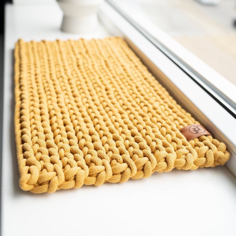 K&ouml;nnte beinhalten: Senfgelber rechteckiger Strickteppich. Der Teppich besteht aus dickem, geknotetem Garn, wodurch eine strukturierte Oberfl&auml;che entsteht. Ein kleines braunes Lederetikett mit Logo ist an der Ecke befestigt. Der Teppich liegt auf einer wei&szlig;en Oberfl&auml;che in der N&auml;he eines Fensters.