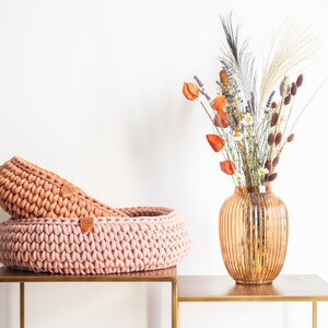 May include: Two handmade, round, knitted baskets in shades of pink and peach sit on a gold-colored table. A decorative vase with dried flowers in orange, white, and brown is to the right.