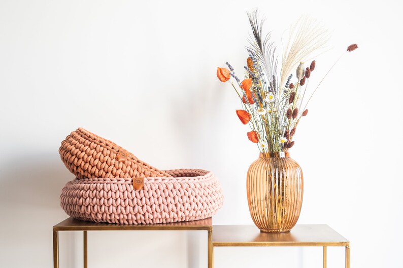 May include: Two handmade, round, knitted baskets in shades of pink and peach sit on a gold-colored table. A decorative vase with dried flowers in orange, white, and brown is to the right.