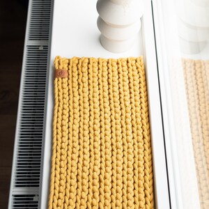 K&ouml;nnte beinhalten: Senfgelber rechteckiger Strickteppich mit Lederetikett. Der Teppich ist aus dickem, grobem Garn gefertigt und liegt auf einer wei&szlig;en Oberfl&auml;che. Im Hintergrund befindet sich eine wei&szlig;e Vase mit Trockenblumen.