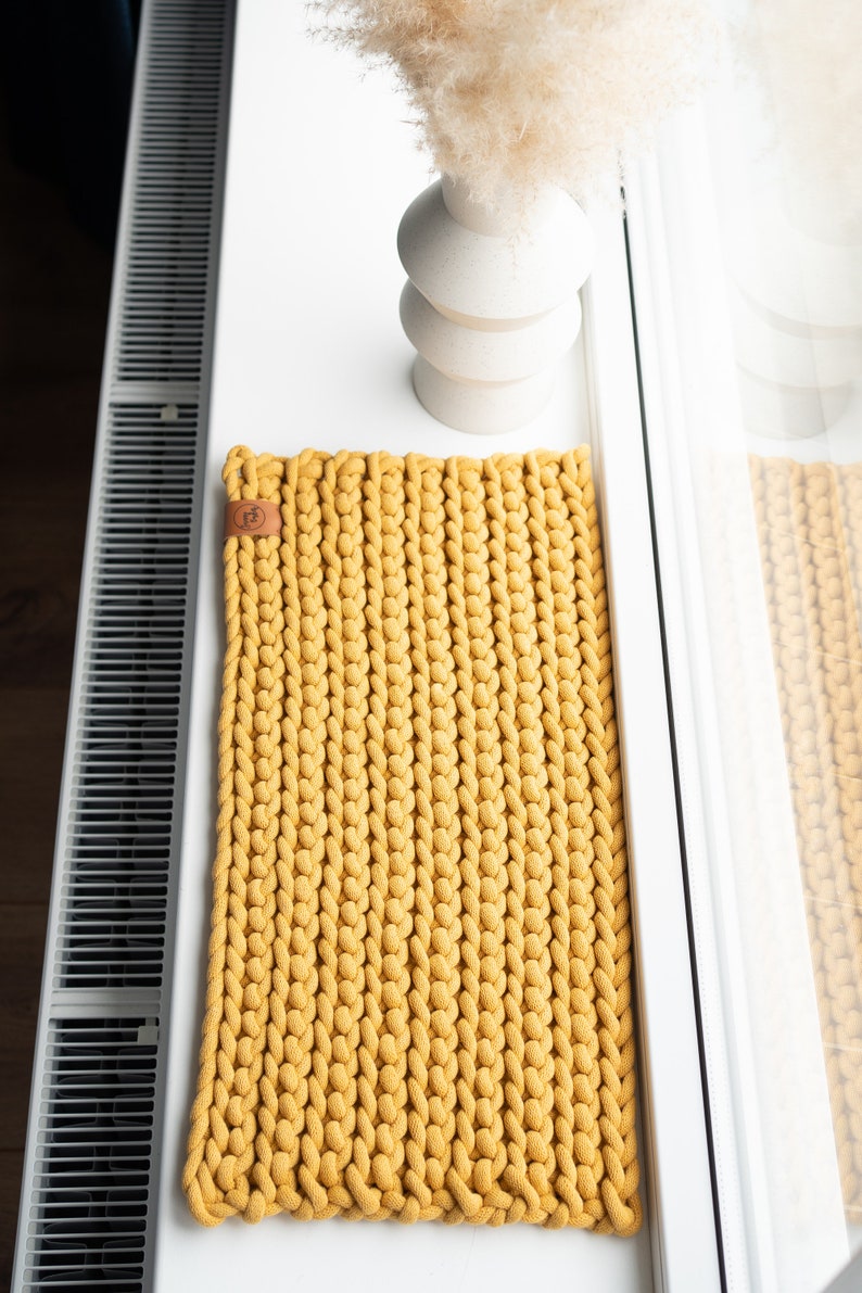 K&ouml;nnte beinhalten: Senfgelber rechteckiger Strickteppich mit Lederetikett. Der Teppich ist aus dickem, grobem Garn gefertigt und liegt auf einer wei&szlig;en Oberfl&auml;che. Im Hintergrund befindet sich eine wei&szlig;e Vase mit Trockenblumen.