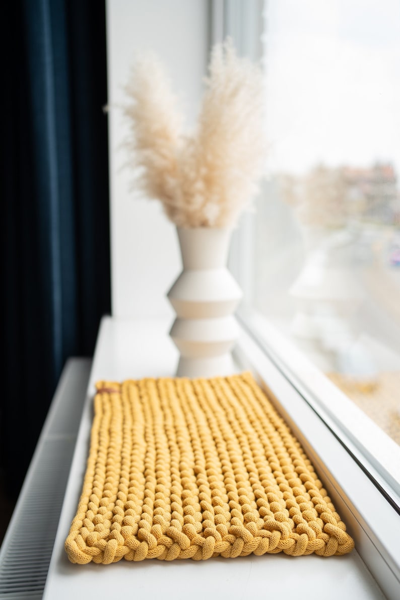 K&ouml;nnte beinhalten: Senfgelber rechteckiger Strickteppich. Der Teppich besteht aus dickem, strukturiertem Garn und hat einen geflochtenen Rand. Im Hintergrund steht eine wei&szlig;e Vase mit getrocknetem Pampasgras. Der Teppich ist ca. 61 cm lang und 41 cm breit.