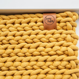 K&ouml;nnte beinhalten: Nahaufnahme eines senfgelben, grob gestrickten Textils. Das Gewebe ist dick und strukturiert, mit einem geflochtenen Muster. Ein braunes Lederetikett mit dem Text "Sunny Bakas" ist am Textil befestigt.