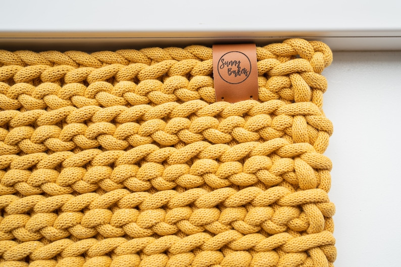 K&ouml;nnte beinhalten: Nahaufnahme eines senfgelben, grob gestrickten Textils. Das Gewebe ist dick und strukturiert, mit einem geflochtenen Muster. Ein braunes Lederetikett mit dem Text "Sunny Bakas" ist am Textil befestigt.