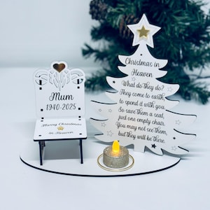 Op de afbeelding: Een witte kerstherdenkingsdecoratie. Het heeft een stoel met de tekst "Mum 1940-2025" en een kerstboom met de tekst "Christmas in Heaven". Een kleine kaars staat voor de boom.