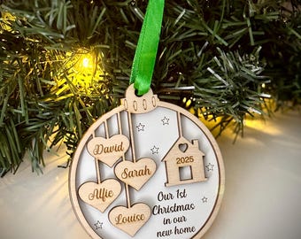 May include: A wooden Christmas ornament with a green ribbon, hanging on a Christmas tree. The ornament has a white background with the names David, Sarah, Alfie, and Louise in heart shapes. It also features a house with the year 2025 and the text "Our 1st Christmas in our new home."