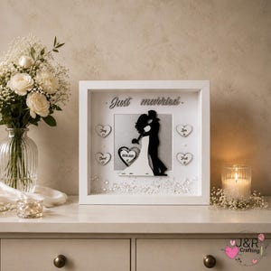 May include: A white square shadow box frame featuring a black silhouette of a couple, the text "Just married," and four small white hearts with names. The display includes a central heart with the date 28.05.2020 and loose clear crystals.