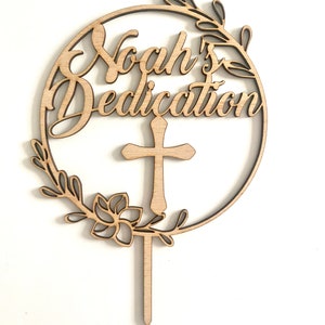 May include: A wooden cake topper featuring the inscription "Noah's Dedication" in a cursive font, encircled by a floral wreath and a cross. Crafted from light-toned wood, it includes a pointed stick for cake insertion.
