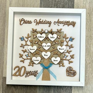 Könnte beinhalten: Ein weißer Rahmen mit einem Holz-Familienbaum im Inneren. Der Baum ist mit blauen und silbernen Verzierungen geschmückt. Der Text "China Wedding Anniversary 20 years" ist am unteren Rand des Rahmens zu sehen.