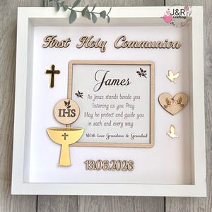 May include: A white framed shadow box with wooden embellishments for a First Holy Communion. The design includes a cross, chalice, doves, and the name "James" with a date. The text reads "First Holy Communion" and a prayer.