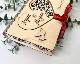 Puede incluir: Una caja de madera con un recorte en forma de corazón y un diseño de paloma. La caja está decorada con una cinta roja y los nombres "Ryan & Megan" y la fecha "29.04.2025" están grabados en la parte delantera.