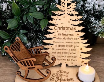 Puede incluir: Un adorno de árbol de Navidad de madera clara con una estrella en la punta y texto grabado sobre la Navidad en el cielo. Una pequeña mecedora de madera teñida de oscuro se asienta sobre la base junto a una vela blanca y el texto Merry Christmas in Heaven Dad.