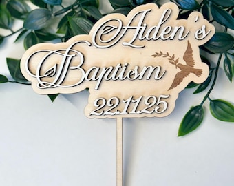 May include: Wooden baptism cake topper with the words "Aiden's Baptism" in a decorative font. A dove carrying an olive branch is etched on the right side. The date "22.11.25" is below the text. The topper has a wooden stick for insertion.