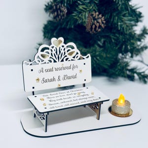 May include: A white decorative bench with the text "A seat reserved for Sarah & David". The bench has a decorative top with gold accents and a small candle holder with a flickering candle. The bench is on a white base.