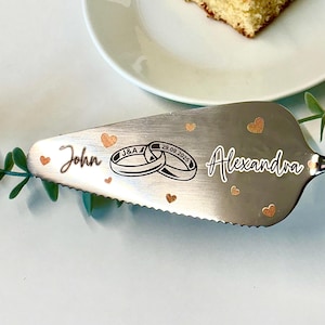 May include: A silver cake server engraved with the names "John" and "Alexandra", the date "29.09.2025", and two wedding rings. It is decorated with small gold hearts. The server is next to a slice of cake.