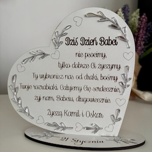 Puede incluir: Un letrero de madera blanco en forma de corazón con un mensaje para una abuela. El letrero dice "Dziś Dzień Babci" que se traduce como "Hoy es el Día de la Abuela" en español. El letrero también incluye el texto "nie psocimy, tylko dobrze Ci życzymy. Ty wybronisz nas od draki, bośmy Twoje rozrabiaki. Całujemy Cię serdecznie, żyj nam, Babciu, długowiecznie. Życzą Kamil i Oskar 21 Stycznia".