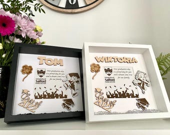 May include: Two white shadow boxes with black frames. The top box has the name "TOM" in wooden letters and the bottom box has the name "WIKTORIA" in wooden letters. Both boxes have a graduation theme with a University of Salford logo, a graduation cap, and a silhouette of a crowd of people.
