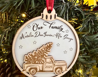 May include: Wooden Christmas ornament with a red ribbon. The ornament features the text "Our Family" and names. A truck carrying a Christmas tree is also depicted, with the year 2025. The ornament is hanging on a Christmas tree.