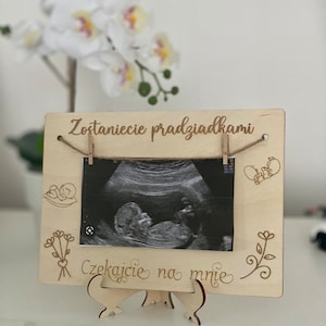 Może przedstawiać: Drewniana ramka na zdjęcia z obrazem USG dziecka. Ramka ma dekoracyjne grawerunki i polski tekst "Zostaniecie pradziadkami" i "Czekajcie na mnie". Jest wspierana przez drewnianą podstawkę.
