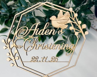 May include: A wooden cake topper with the words "Aiden's Christening" and the date 22.11.25. The topper features a dove and floral accents within a geometric frame. The topper is made of light-colored wood.