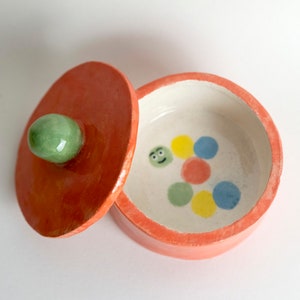 Puede incluir: Una pequeña caja de cerámica naranja con un pomo verde en la tapa. El interior de la caja es blanco con un diseño pintado a mano de una oruga verde sonriente con puntos de colores.