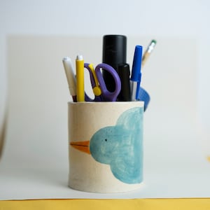 Puede incluir: Un portalápices cilíndrico con un diseño de pájaro azul pintado a mano. El portalápices está lleno de bolígrafos, lápices y tijeras. El pájaro tiene un cuerpo azul y un pico naranja. El portalápices es de un material beige claro.