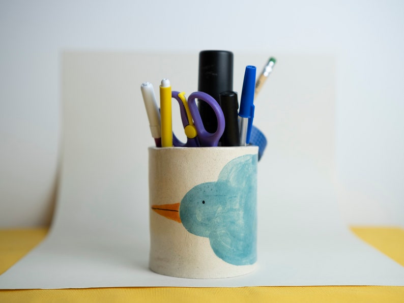 a handmade ceramic pencil holder with an illustrated turquoise blue bird that goes around the whole surface of the pencil holder. It has pencil, pens and markers inside to show the utility side of the object.