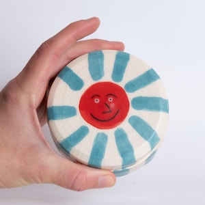 Könnte beinhalten: Ein runder Keramikbehälter mit Deckel, verziert mit einem fröhlichen Sonnenmotiv. Die Sonne hat ein rotes Gesicht mit einem lächelnden Ausdruck, umgeben von blauen Strahlen auf cremefarbenem Hintergrund. Der Behälter wird in einer Hand gehalten.