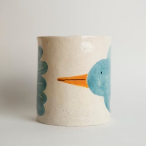 the bird pencil cup in a white background. You can see the ceramic piece has glaze and it has a beige stoneware raw look. The bird illustration shows up in one of the sides, you can see both beak and tail of the bird. the beak is orange.