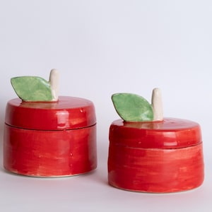Puede incluir: Dos recipientes de cerámica con forma de manzana. Los recipientes son de un rojo vibrante con un acabado brillante. Cada uno tiene una hoja verde y un tallo blanco en la parte superior. Los recipientes están diseñados para parecerse a manzanas.