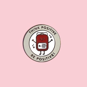 May include: A round pin with a white background and a red cartoon blood bag with a happy face. The blood bag has a label that says "B+". The pin has a gray border with the text "Think Positive" and "Be Positive!" around the edge.